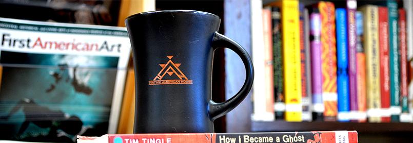 Picture of a coffee mug that says "Native American House" with a bookshelf full of books in the background.